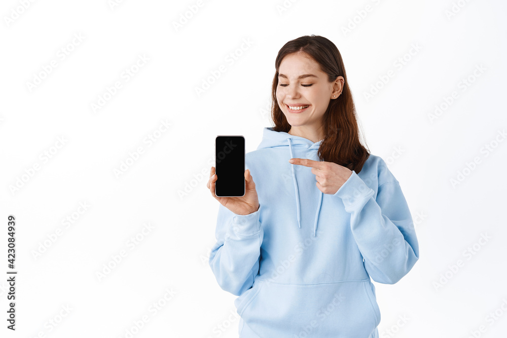 Beautiful teenage girl student pointing and showing empty smartphone screen, mobile application, smiling pleased, standing against white background Beautiful teenage girl student pointing and showing empty smartphone screen, mobile application, smiling pleased, standing against white background