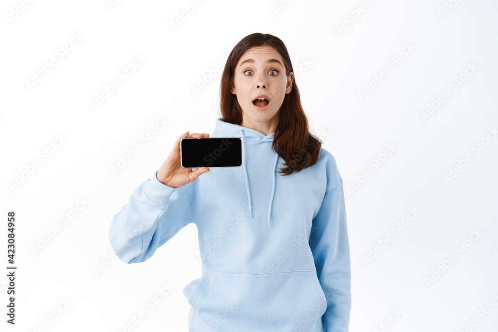 Excited teenage girl showing application or video game on empty mobile phone screen, demonstrates your promo on smartphone display, standing against white background