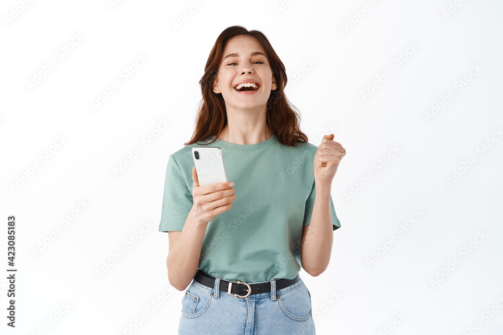 Young happy woman say yes, winning prize or daily goal on mobile app, holding smartphone and rejoicing, celebrating victory in application, standing against white background Young happy woman say yes, winning prize or daily goal on mobile app, holding smartphone and rejoicing, celebrating victory in application, standing against white background