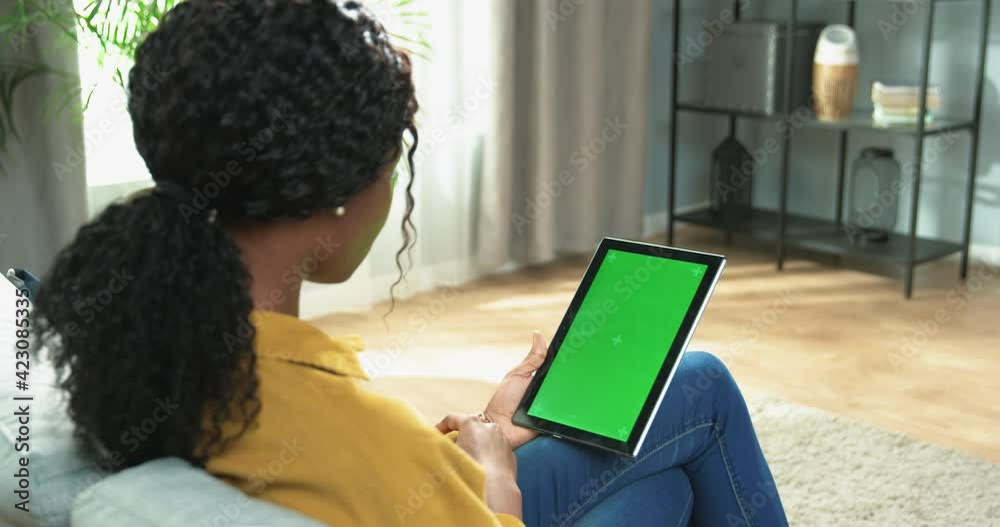 Over shoulder view of African American woman sitting on sofa in apartment in living room tapping on tablet device with green screen with tracking motion browsing online, technology concept