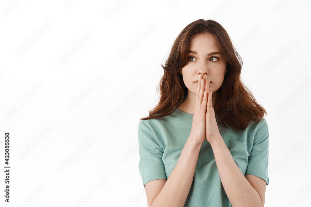 Young hopeful woman praying, looking aside with worried face, pleading everything be okay, concerned for someone, standing over white background Young hopeful woman praying, looking aside with worried face, pleading everything be okay, concerned for someone, standing over white background