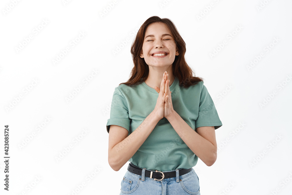 Happy young woman feeling grateful and delighted, holding hands in pray namaste sign, close eyes and smiling broadly, thankful for opportunity, standing against white background Happy young woman feeling grateful and delighted, holding hands in pray namaste sign, close eyes and smiling broadly, thankful for opportunity, standing against white background