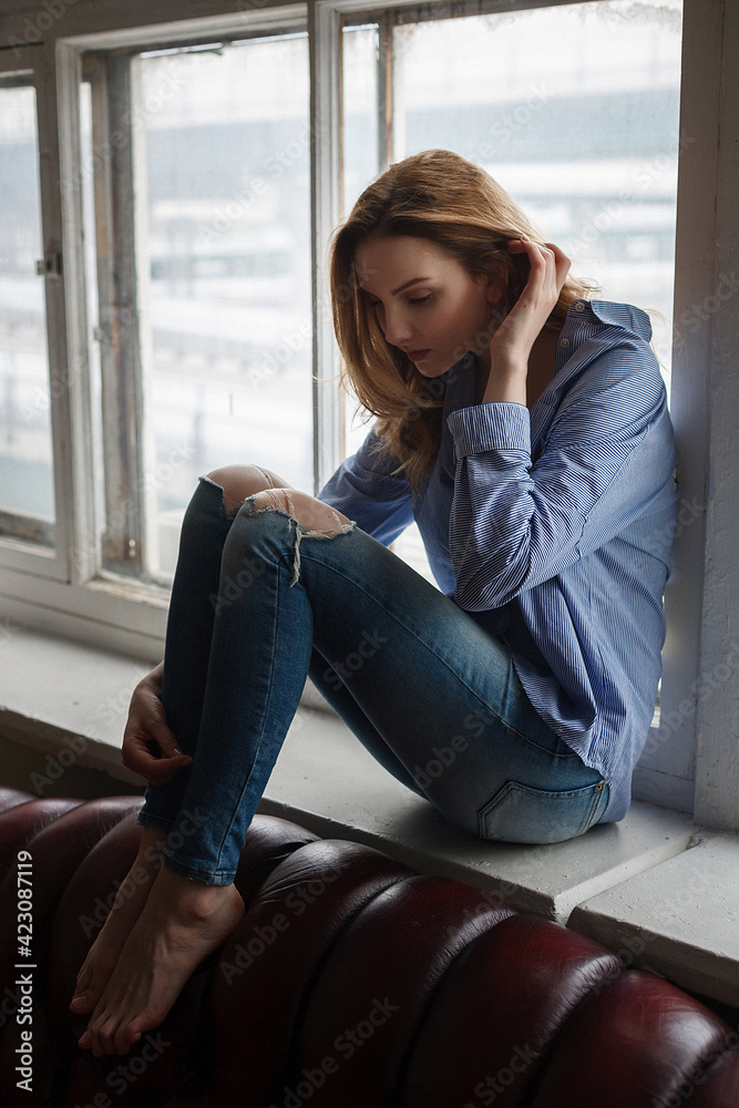 young woman wearing blue striped shirt and ripped jeans sitting on windowsill young woman wearing blue striped shirt and ripped jeans sitting on windowsill