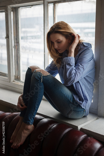 young woman wearing blue striped shirt and ripped jeans sitting on windowsill