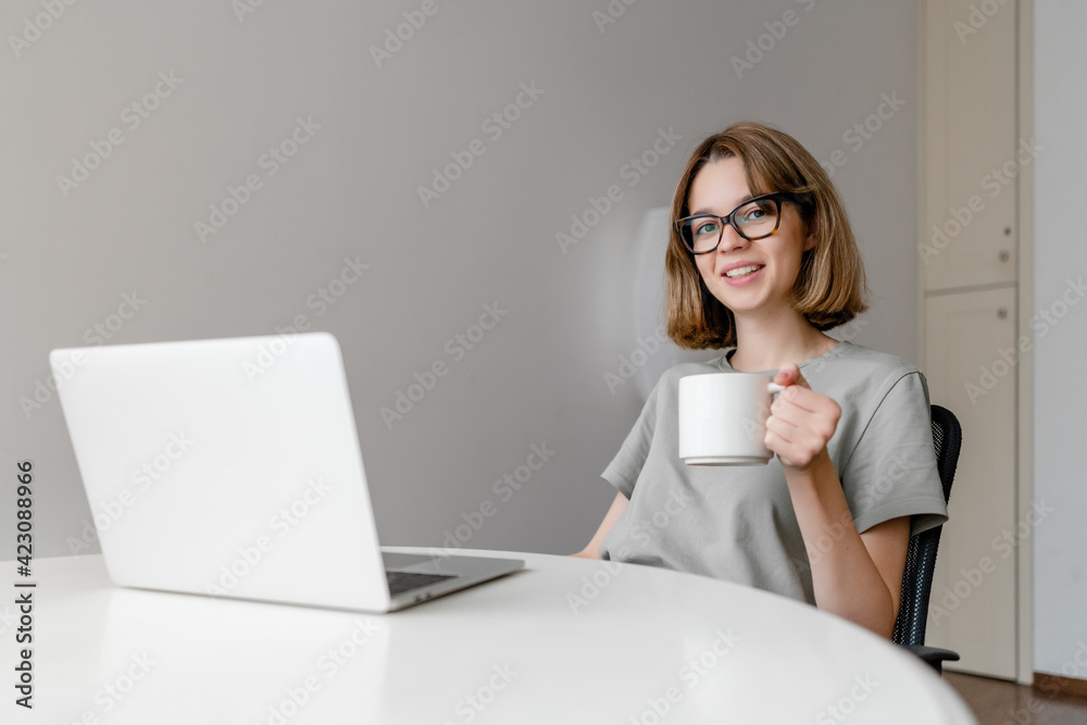 young relaxed caucasian woman smiling sitting with laptop in the apartment with cup in her hands young relaxed caucasian woman smiling sitting with laptop in the apartment with cup in her hands