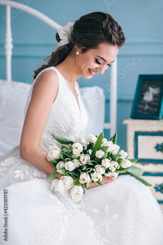 Beautiful bride with a wedding bouquet sitting on bed indoors in studio interior like at home. photo