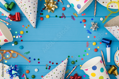 Top view photo of birthday party composition spiral cocktail tubes ribbon stars candles pipes hats confetti paper cups and plates on isolated blue wooden table background with copyspace in the middle