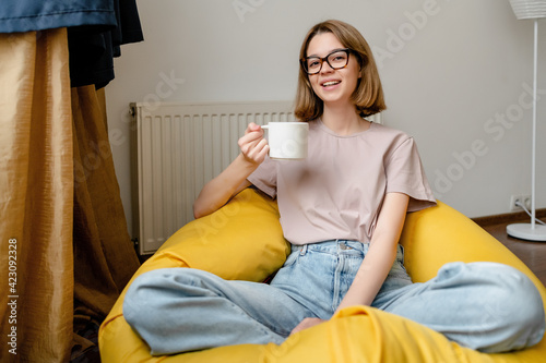young smiling woman sitting in the apartment wearing blue jeans pink tshirt and glasses holding a cup
