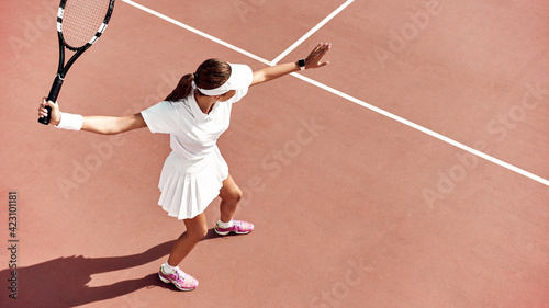 Pretty girl plays tennis on the court outdoors. She prepares to beat on a ball. Woman wears a light blue sportswear with white sneakers. Top view photo. © Friends Stock