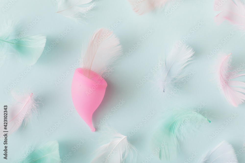 Fototapeta premium Menstrual cup and feathers on blue background. Top view, flat lay