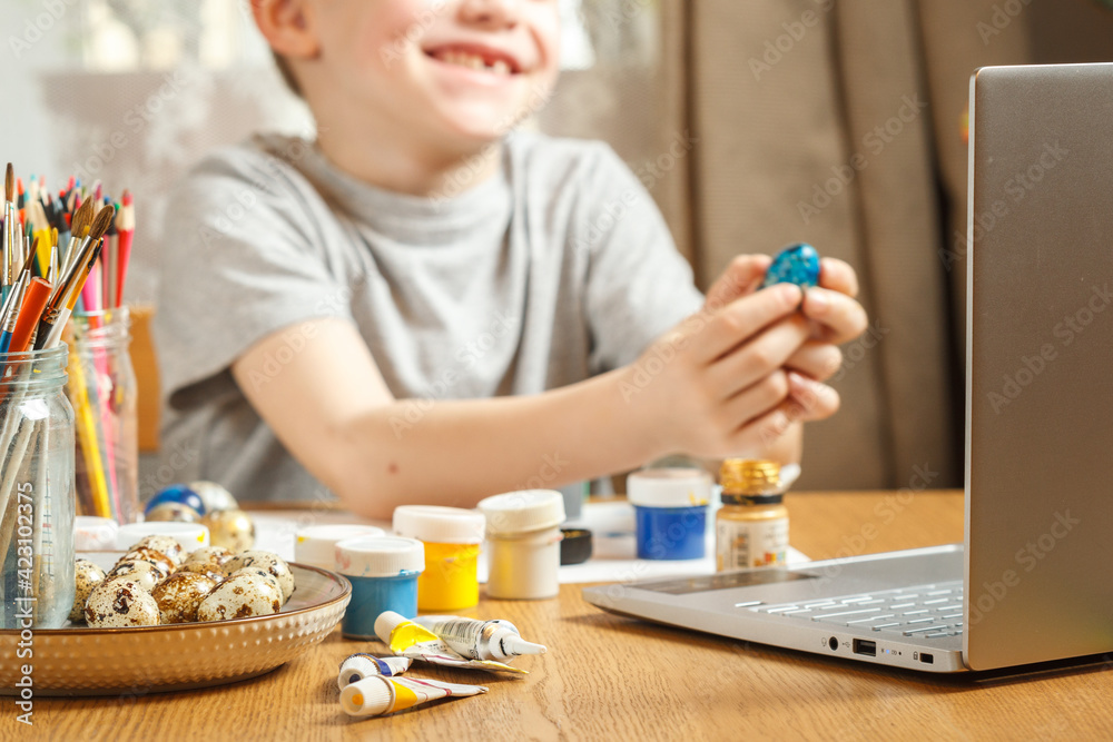 Easter greetings online. A smiling child shows a colored Easter egg, a video call to friends, relatives. Focus on the laptop. Blurred background Easter greetings online. A smiling child shows a colored Easter egg, a video call to friends, relatives. Focus on the laptop. Blurred background
