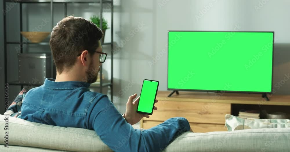 Caucasian man in glasses sitting on sofa in modern room in house watching TV with green screen and typing on smartphone with chroma key browsing online. Back view, leisure concept