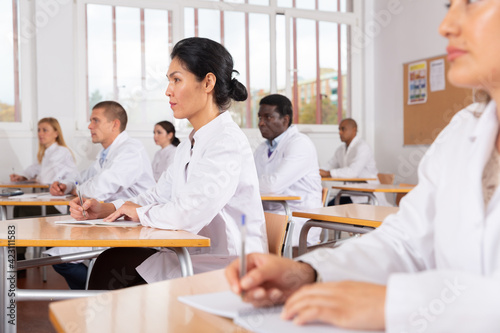 Young positive woman in white medical coat sitting at desk in classroom attending seminar