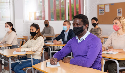 Portrait of confident young adult male in face mask for viral protection studying in classroom with colleagues