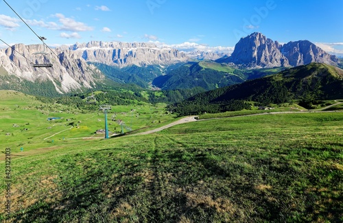 Summer scenery of a beautiful valley viewed from Seceda, with rugged Sella & Sassolungo-Sassopiatto Mountains in the background & a chairlift gliding over green meadows in Col Raiser, Dolomites, Italy