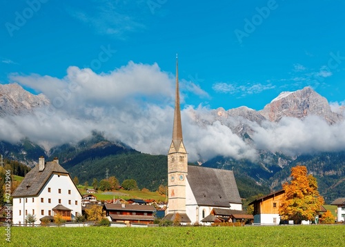 A peaceful village at the foothills of Steinernes Meer Mountains in Maria Alm, Austria, with a majestic church tower (Wallfahrtskirche) standing tall in green fields & foggy mountains in background photo