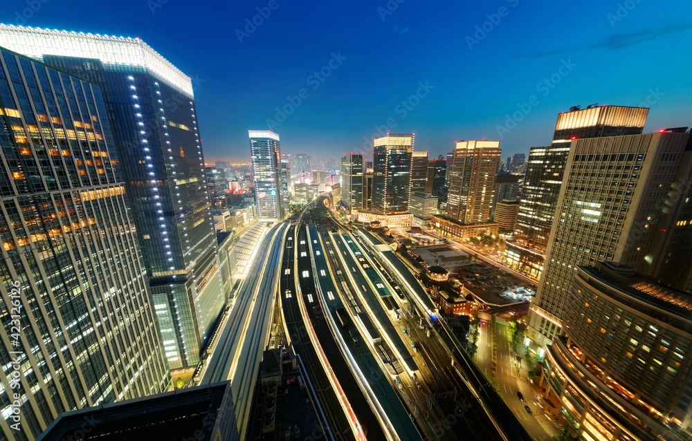 Obraz premium Beautiful nightscape of Tokyo Train Station with a high angle view of railway platforms stretching between modern office towers & city lights glistening under blue evening sky in Chiyoda, Tokyo, Japan