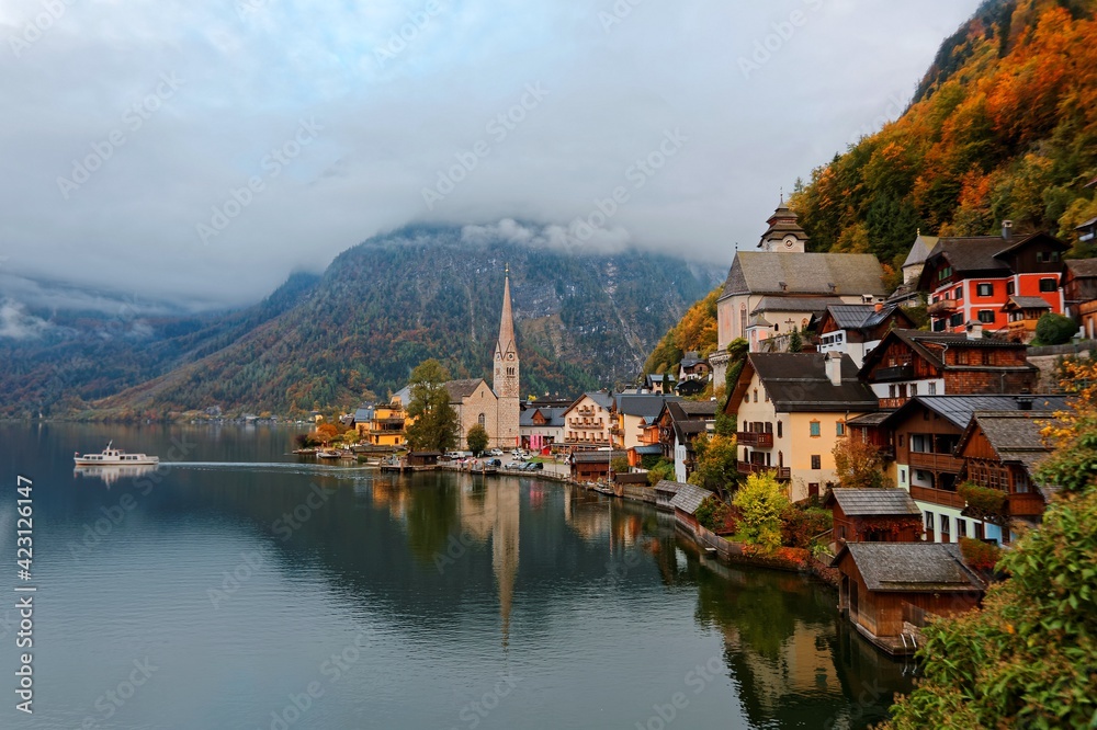 Morning scenery of Hallstatt, a lakeside village in Salzkammergut area of Austria, with a tourist boat cruising on the peaceful lake and foggy alpine mountains in background in colorful autumn season Morning scenery of Hallstatt, a lakeside village in Salzkammergut area of Austria, with a tourist boat cruising on the peaceful lake and foggy alpine mountains in background in colorful autumn season