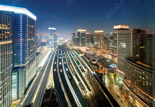 Beautiful nightscape of Tokyo Train Station with a high angle view of railway platforms stretching between modern office towers & city lights glistening under blue evening sky in Chiyoda, Tokyo, Japan