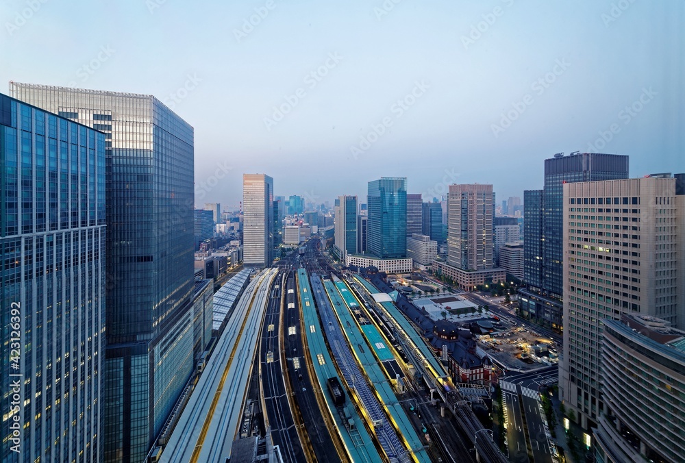 Scenery of Tokyo Train Station before dawn, with a high angle view of railway platforms stretching out between modern office towers & city lights glistening in blue twilight in Chiyoda, Tokyo, Japan