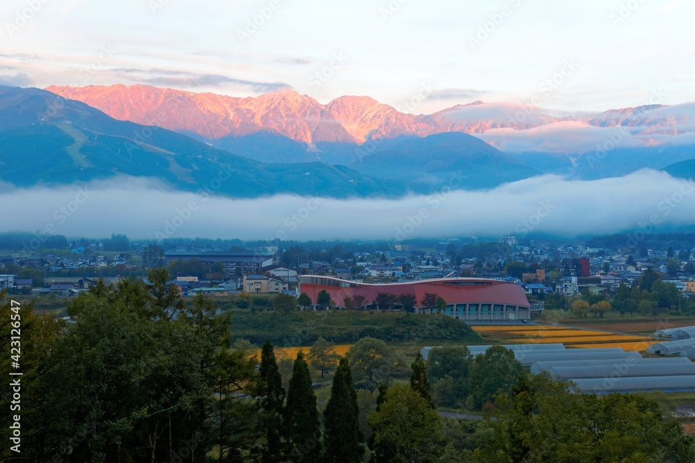 Sunrise scenery of majestic Hakuba Mountain Range with golden glow on the submit, a fog belt on the mountainside and village houses in the field on a foggy autumn morning in Nagano, Japan