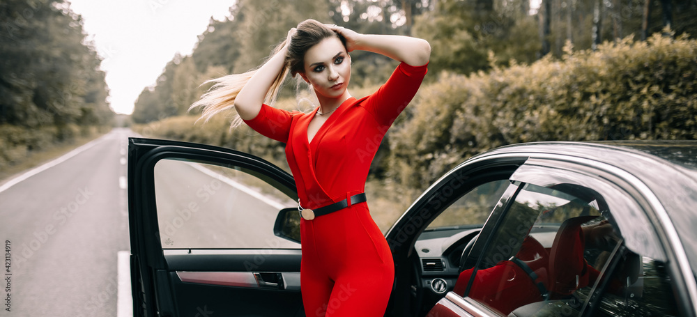 A beautiful young girl in a red overalls stands by a black car on an empty road in the forest A beautiful young girl in a red overalls stands by a black car on an empty road in the forest