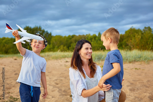 A young happy mother with two boys playing on the sand near the forest, in the hands of one of them a model of a civil aircraft and a hat on his head
