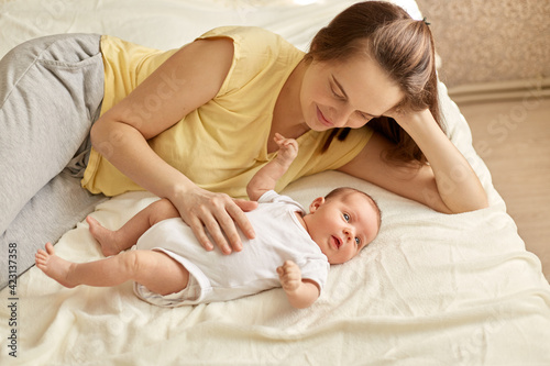Happy mother lying on bed on blanket and looking at her little newborn baby and touching belly with love and gentle, infant wearing white bodysuit, family posing indoor.