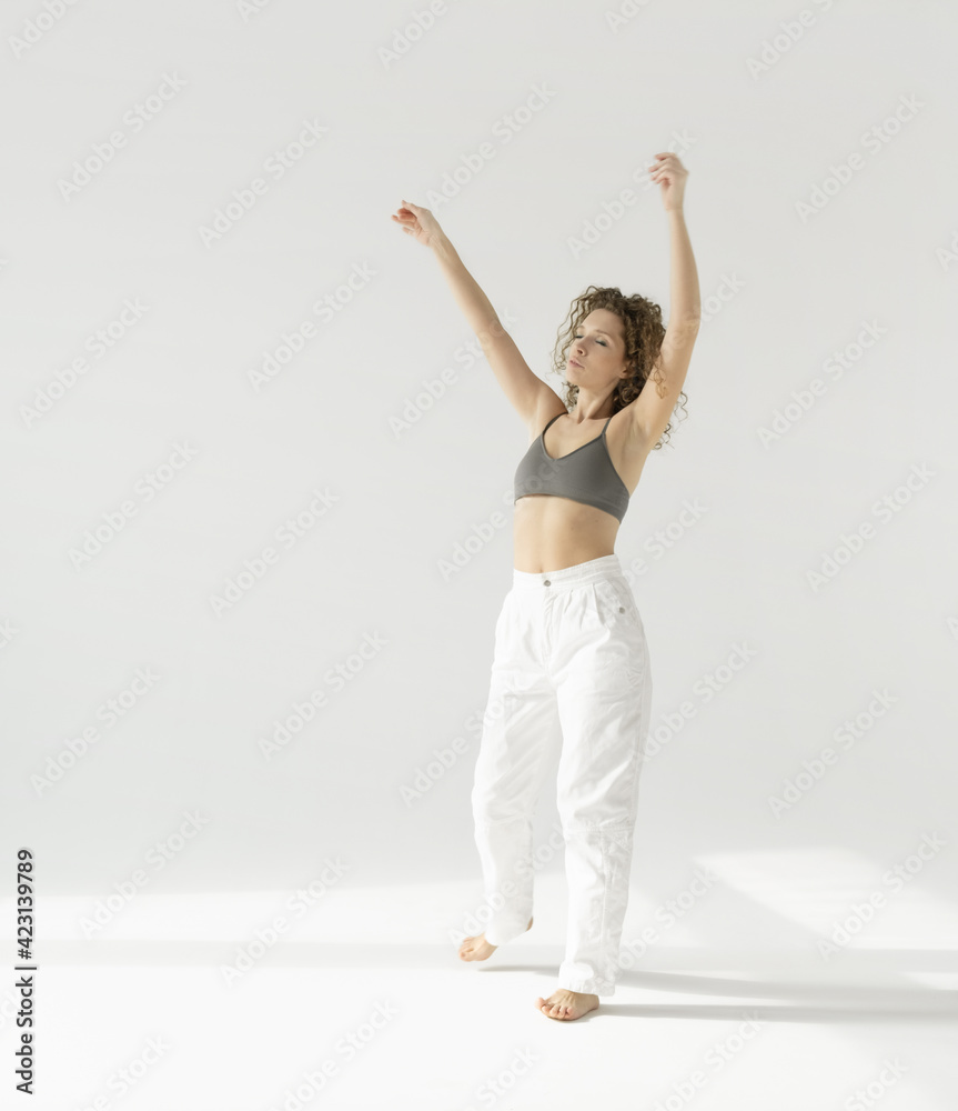 a European woman in white trousers and a top slowly dances against a white background lit by sunlight. Natural full-length portrait of a young woman moving in the morning, her hands raised in the air