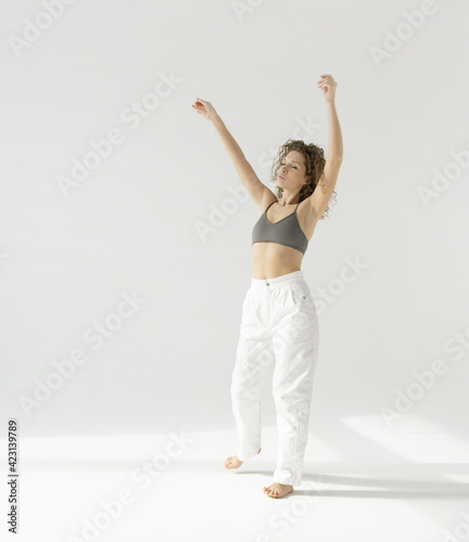 a European woman in white trousers and a top slowly dances against a white background lit by sunlight. Natural full-length portrait of a young woman moving in the morning, her hands raised in the air