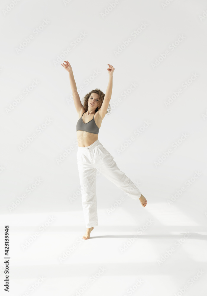 Fototapeta premium a European woman in white trousers and a top slowly dances against a white background lit by sunlight. Natural full-length portrait of a young woman moving in the morning, her hands raised in the air