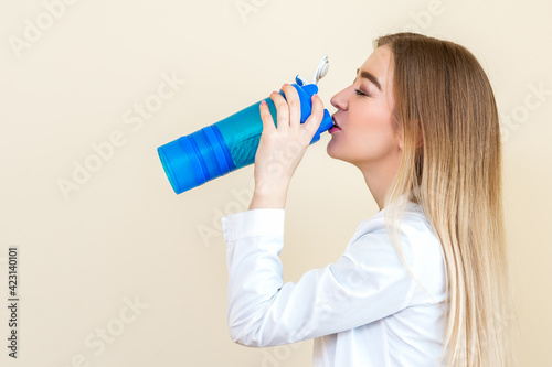 Side view of beautiful young caucasian woman is drinking water from plastic bottle against a beige background
