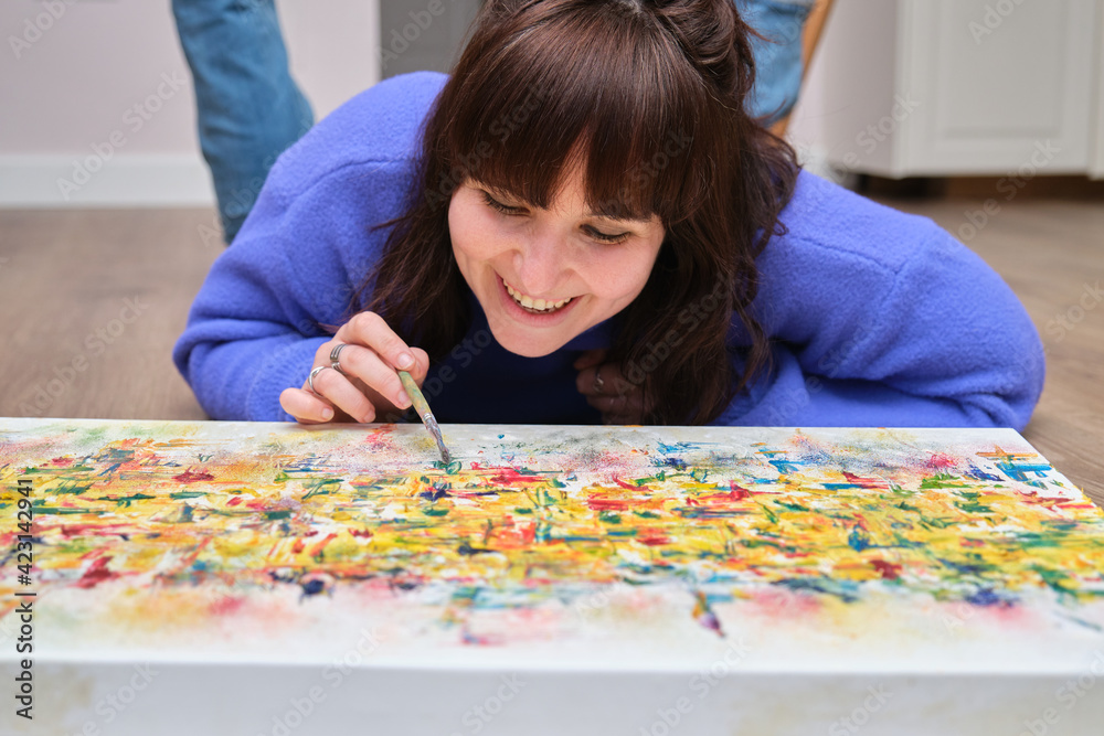 Young woman laughing and painting on canvas with oil paints at home. Artist drawing laying on the floor. Young woman laughing and painting on canvas with oil paints at home. Artist drawing laying on the floor.