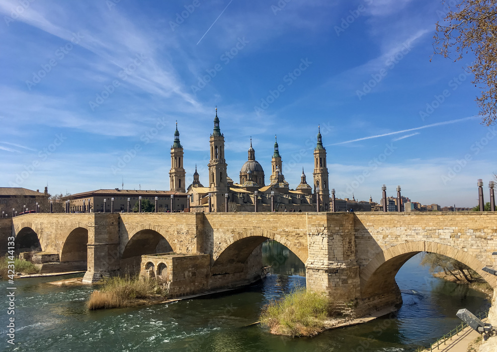 People cross the river Ebro on the stone bridge with the Cathedral of Zaragoza in the background.
