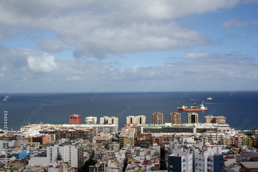 High view of the city of Las Palmas de Gran Canaria with the Atlantic Ocean in the background, with buildings in the foreground and some boats in the sea. Calm ocean creates a straight horizon line.