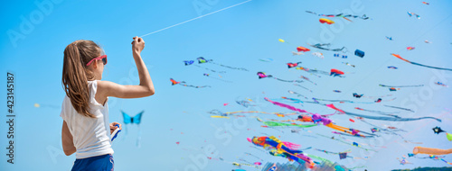 The little girl in the t-shirt plays on the beach by the sea with a colorful kite. In the background many people are walking on the beach.