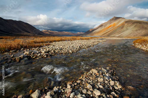 Autumn Arctic landscape. View of a small river in the tundra. In the distance, a lake and mountains. The slope of the hill is an unusual orange color. Chukotka, Polar Siberia, Far North of Russia.