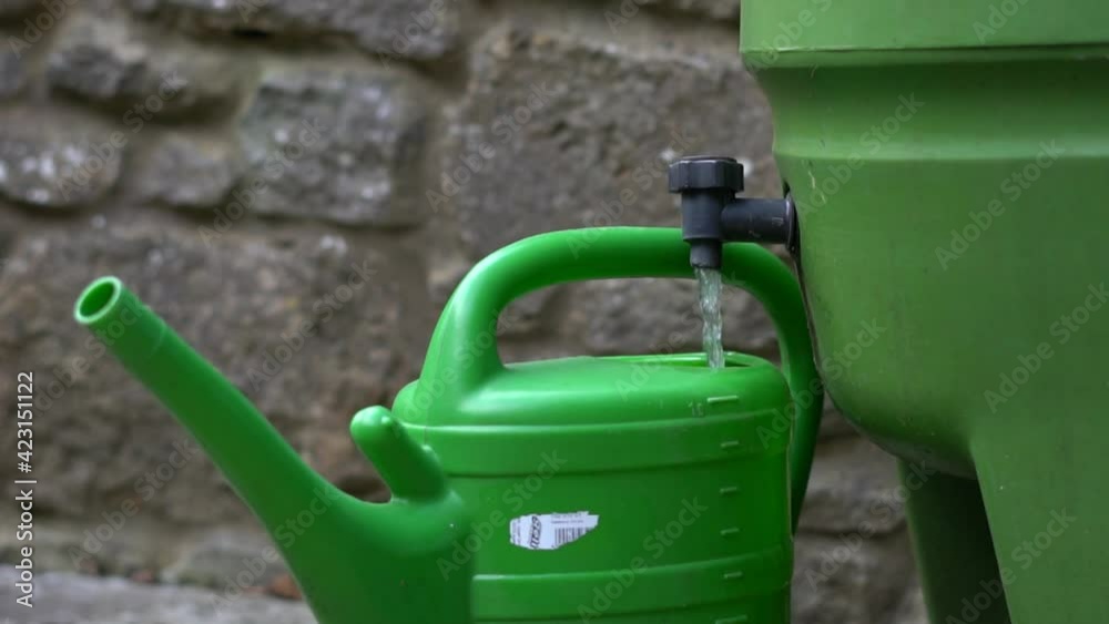 Green watering can being filled via outside tap on the side of a rain water filled container. Backyard gardening concept. Green watering can being filled via outside tap on the side of a rain water filled container. Backyard gardening concept.