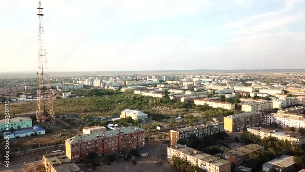A small town on the shore of the Balkhash lake. Low-rise Soviet-built houses. In the center of the city you can see a TV tower and a factory with pipes in the distance. Green trees and bushes grow.