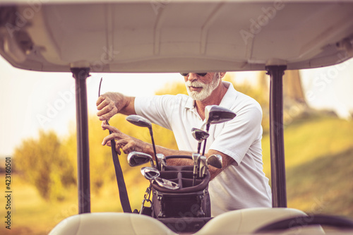 Senior golfer man on golf court packing golf sticks in golf bag.