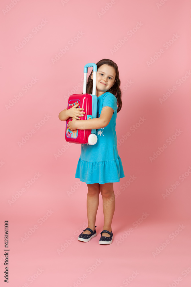 full-length portrait of a long-haired girl hugging a red plastic toy suitcase while waiting for a vacation, isolated on a pink background. full-length portrait of a long-haired girl hugging a red plastic toy suitcase while waiting for a vacation, isolated on a pink background.
