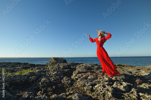 Lovely young slender caucasian woman in a silk pancake dress and a straw hat stands in a picturesque boroco pose on the rocky ocean coast on a sunny morning photo