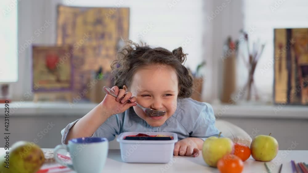 Small schoolgirl eating or having lunch concept. Girl eating her food with fork and laughing while eating. Happy smiling small girl is having dinner at the table. 4k footage Small schoolgirl eating or having lunch concept. Girl eating her food with fork and laughing while eating. Happy smiling small girl is having dinner at the table. 4k footage