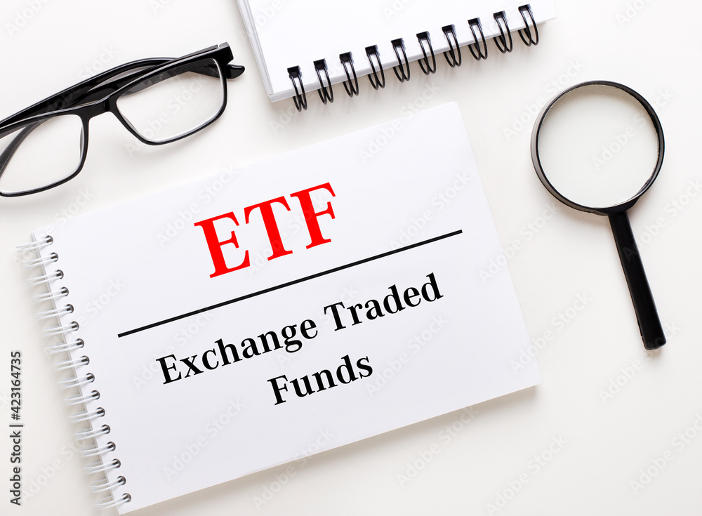 ETF Exchange Traded Funds is written in a white notebook on a light background near the notebook, black-framed glasses and a magnifying glass. ETF Exchange Traded Funds is written in a white notebook on a light background near the notebook, black-framed glasses and a magnifying glass.