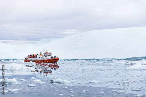 A red boat with people in swimming on the cold sea with many floating icebergs around and with glacier behind, in Greenland,
