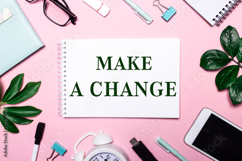 MAKE A CHANGE is written in a white notebook on a pink background surrounded by business accessories and green leaves.