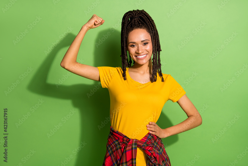 Photo of sweet sporty dark skin lady dressed yellow t-shirt rising fists showing muscles isolated green color background Photo of sweet sporty dark skin lady dressed yellow t-shirt rising fists showing muscles isolated green color background