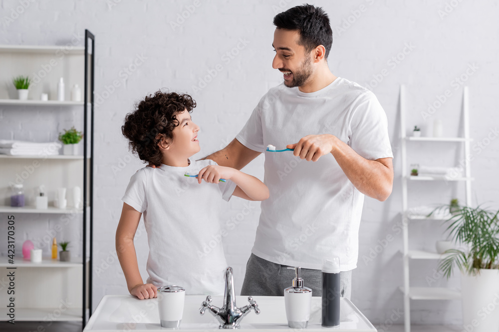 Fototapeta premium Young arabian man looking at son with toothbrush in bathroom