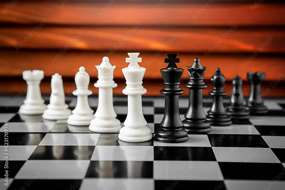 White and black chess pieces stand on a battle chessboard on a red wooden background