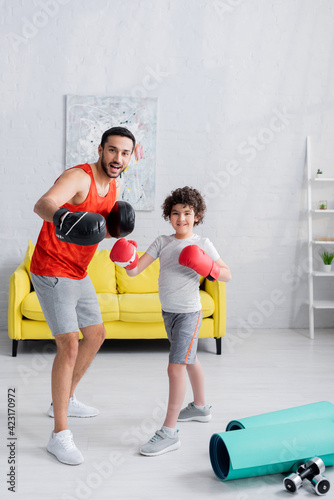 Smiling arabian father and son in boxing gloves smiling at camera near sport equipment at home